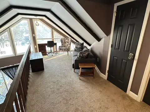 Carpeted loft with angled beamed ceiling, gray sofa, accent chair and desk beneath bright picture windows