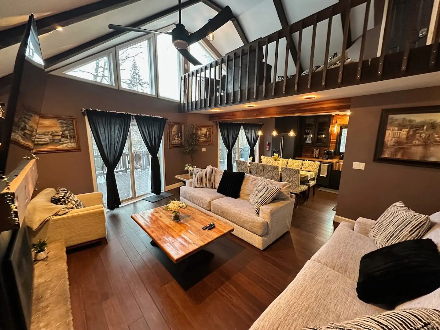 Vaulted great room with loft balcony, wall of windows, cozy sofas, and open flow to the kitchen and dining area