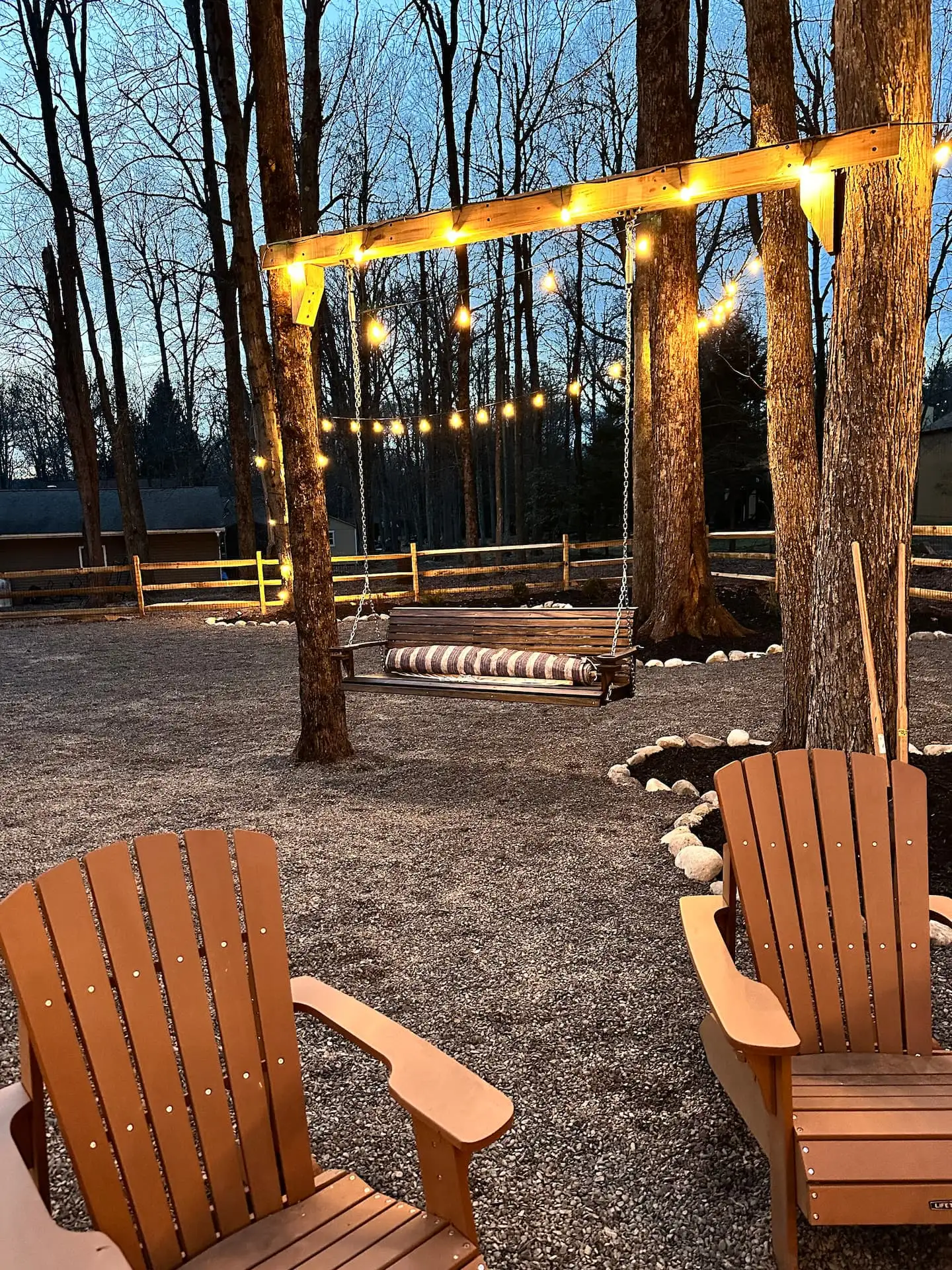 Backyard porch swing under glowing string lights at dusk, framed by tall trees and Adirondack chairs