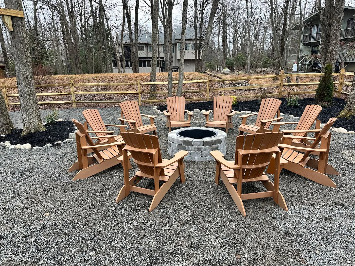 Ten Adirondack chairs circling a stone fire pit on the gravel backyard patio, framed by tall Poconos trees