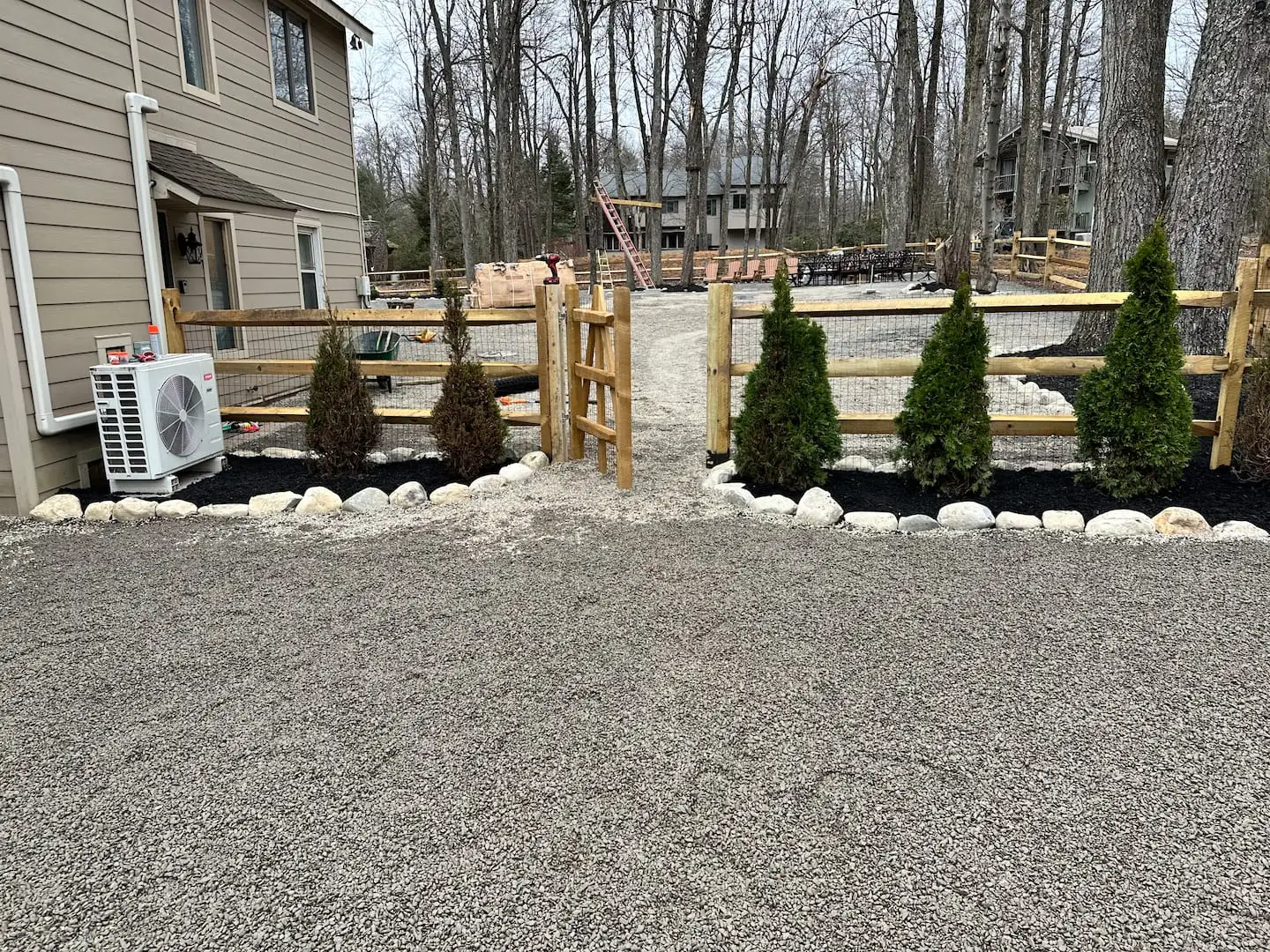 Gated split-rail fence with evergreen shrubs and river-rock borders leading into the graveled backyard