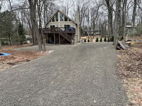 Wide gravel driveway leading to the chalet's peaked front windows, elevated deck, and split-rail fenced yard
