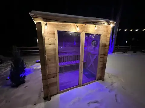 Outdoor cedar sauna cabin lit by Edison string lights and purple glow on a snowy winter night