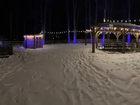 Snowy backyard at night with string lights, glowing gazebo dining area, sauna cabin and hot tub
