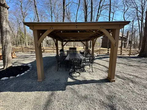 Timber-frame gazebo sheltering a long iron dining table, chairs and grill on the gravel patio