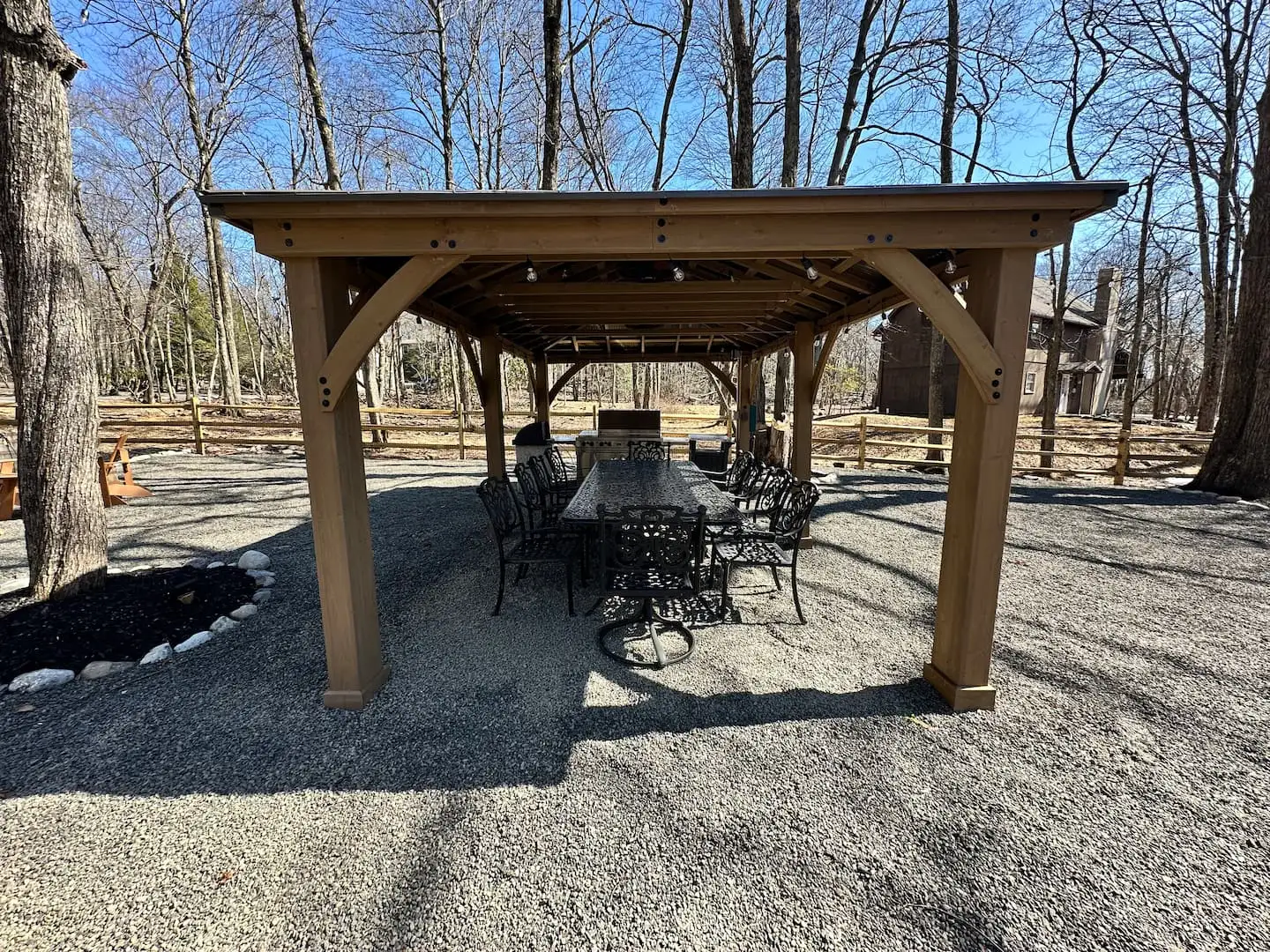 Timber-frame gazebo sheltering a long iron dining table, chairs and grill on the gravel patio