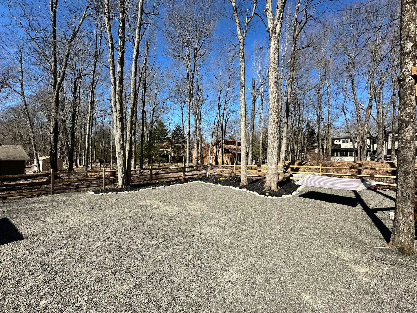 Spacious gravel backyard framed by tall trees, with a hammock, split-rail fence, and stone-lined planting beds