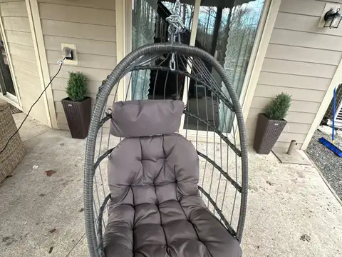 Hanging rattan egg chair with plush cushions on the covered front patio, framed by potted evergreens