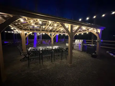 Evening view under the timber pavilion with string lights, wrought-iron dining set, and built-in grill station