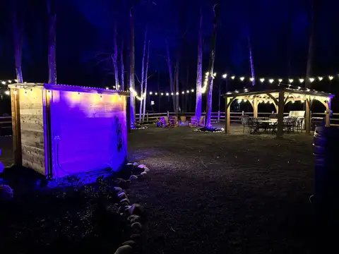Backyard at night with purple-lit cedar sauna, string lights, and gazebo dining area among the trees