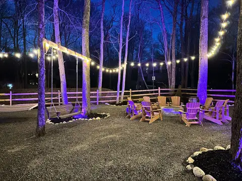 Backyard at dusk with Adirondack chairs around a stone fire pit, porch swing, hammock, and string lights through the trees