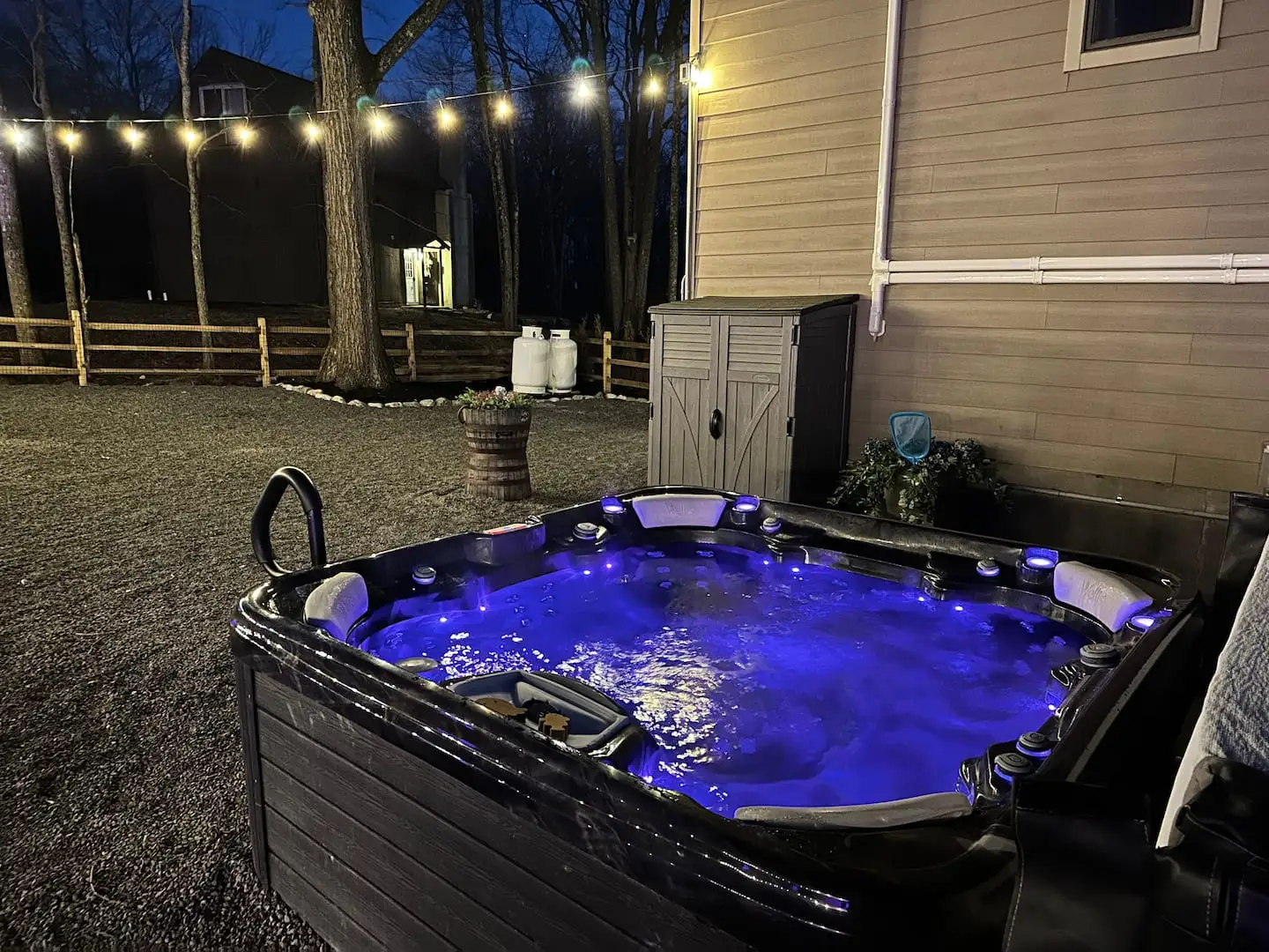 Bubbling hot tub glowing blue at night beside the chalet, with string lights over the fenced backyard