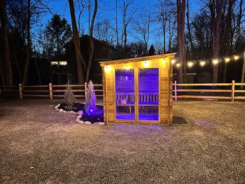 Cedar barrel-style sauna with glass double doors glowing purple at dusk beside a landscaped stone bed