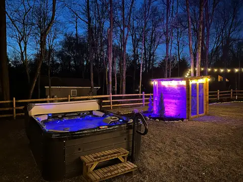 Hot tub steaming at dusk with steps and grab bar, facing the purple-lit barrel sauna under string lights