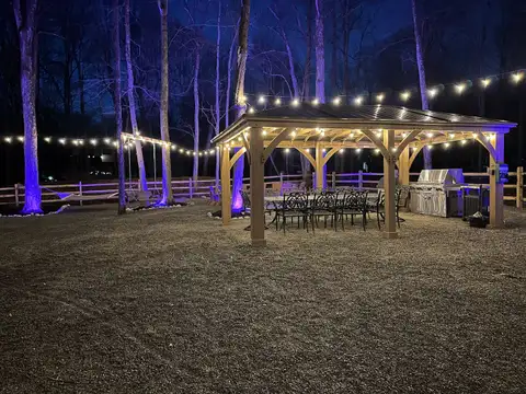 Wooden pavilion glowing with string lights at night, sheltering a long dining table and stainless grill